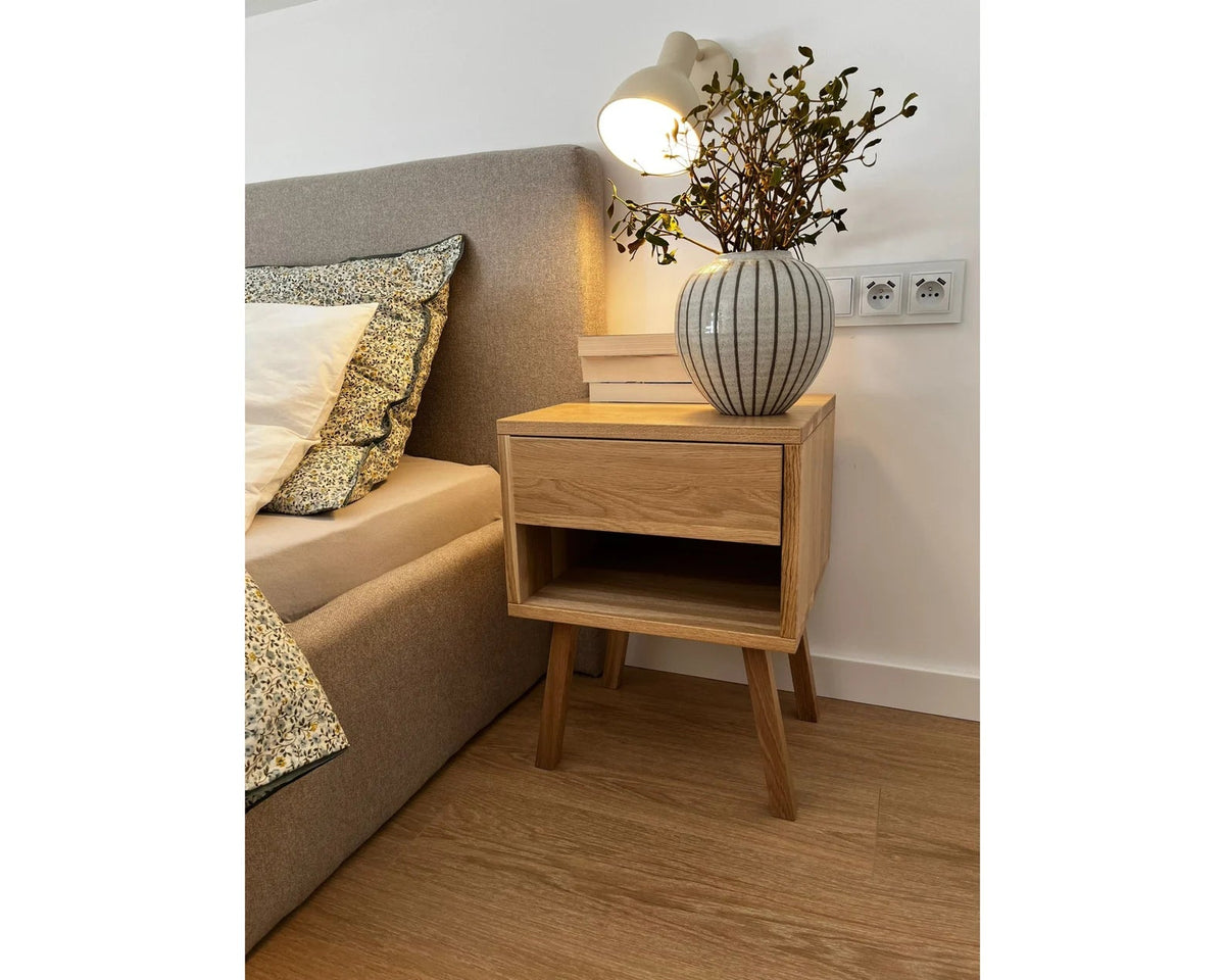 Oak nightstand with open shelf and ceramic vase beside upholstered bed with wall light